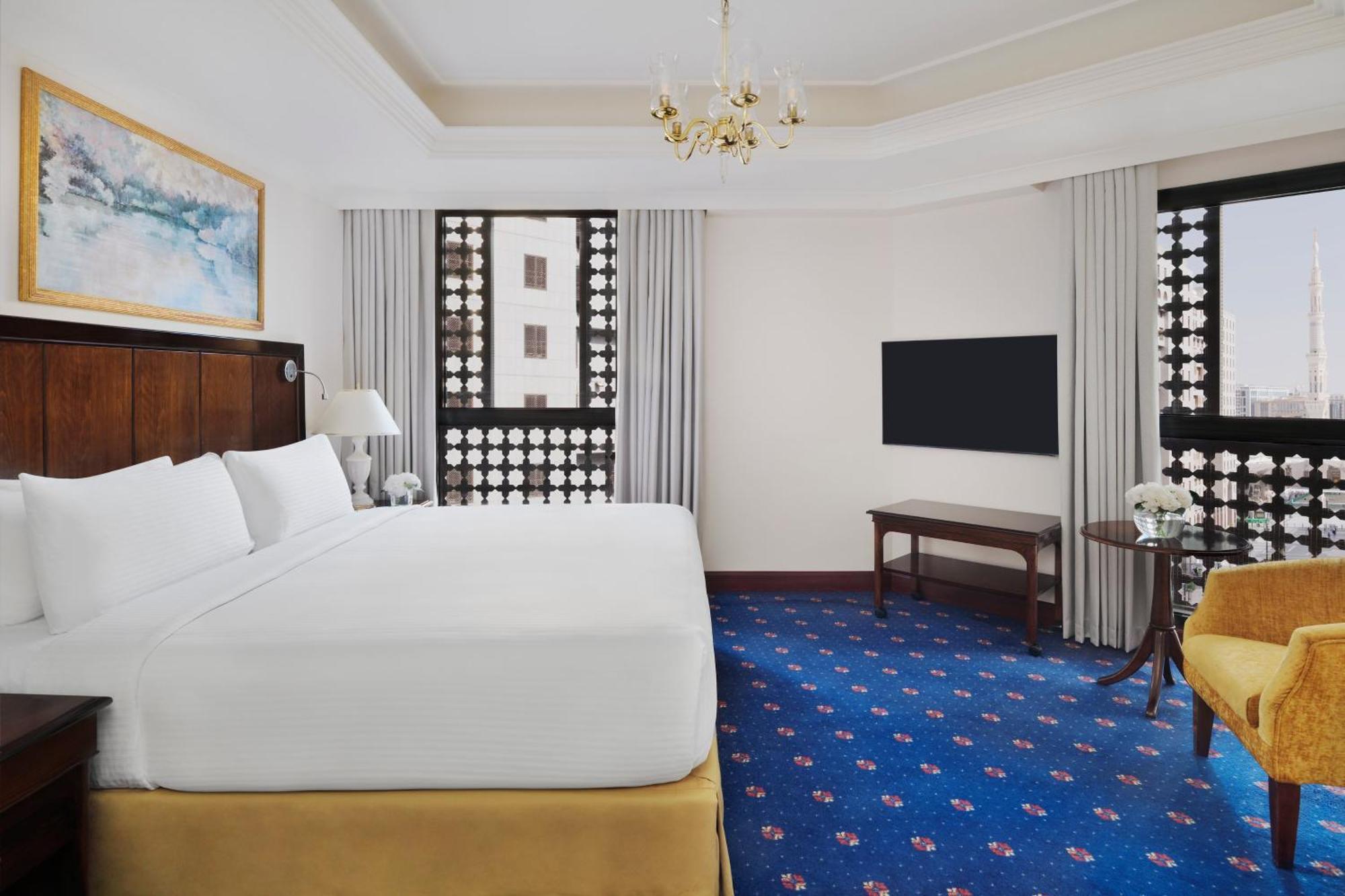 ダー アル ヒジラ インターコンチネンタル マディーナ エクステリア 写真 The photo shows an elegant hotel room. It features a large bed with a white comforter, wooden furniture, and a television mounted on the wall. There are decorative windows with intricate designs that allow natural light to filter in. The floor is cov