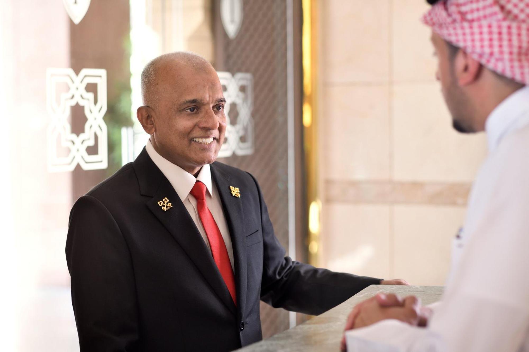 ダー アル ヒジラ インターコンチネンタル マディーナ エクステリア 写真 The photo shows two men engaged in conversation at a hotel reception desk. One man is wearing a dark suit with a red tie and has a welcoming smile. The other man is dressed in traditional attire, possibly a thobe, and a headscarf. The setting appears