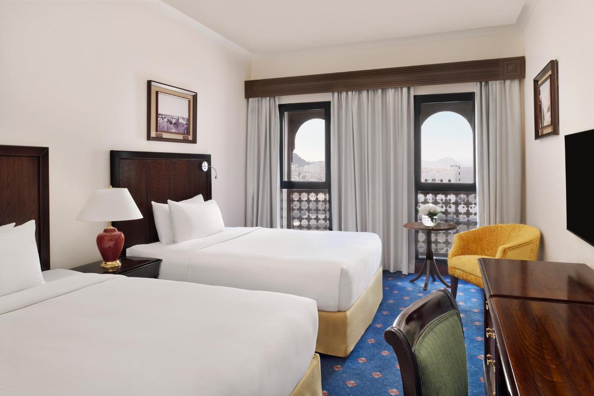 ダー アル ヒジラ インターコンチネンタル マディーナ エクステリア 写真 The photo shows a hotel room featuring two queen-sized beds, each with white bedding. There is a bedside lamp on a small table between the beds. The room has large windows with sheer curtains, allowing natural light to enter. A cozy yellow armchair i