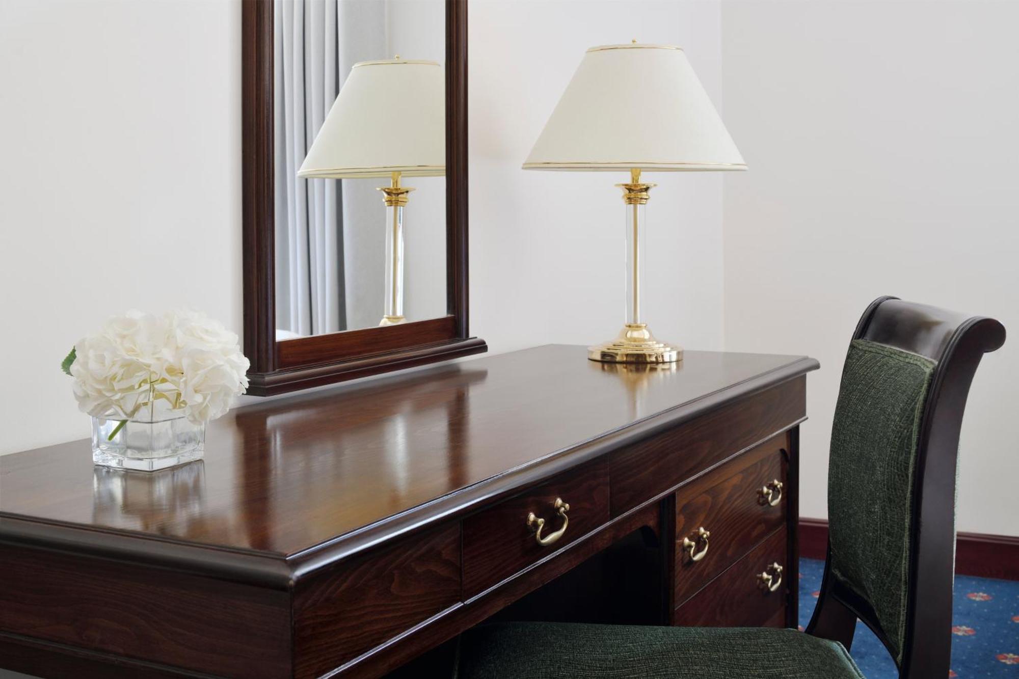 ダー アル ヒジラ インターコンチネンタル マディーナ エクステリア 写真 The photo shows a wooden vanity or desk with a polished surface. It features a large mirror mounted on the wall behind it. On the desk, there is a gold-colored lamp with a white lampshade, and a small glass vase holding white flowers. To the side of 