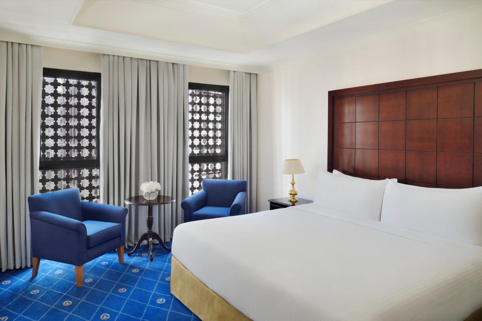 ダー アル ヒジラ インターコンチネンタル マディーナ エクステリア 写真 The photo shows a hotel room featuring a large bed with white bedding. There are two blue armchairs positioned near a small round table, which has a decorative item on it. The room has large windows covered with sheer curtains, allowing natural light