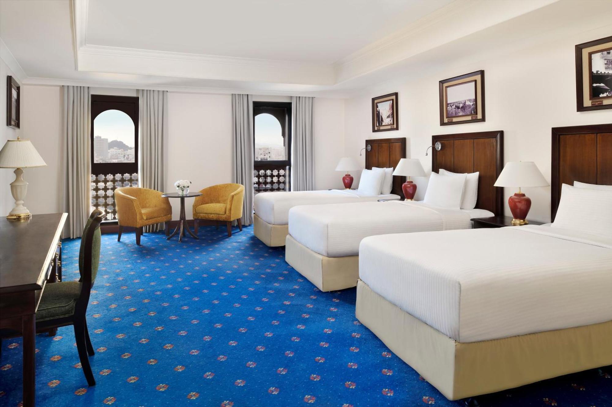 ダー アル ヒジラ インターコンチネンタル マディーナ エクステリア 写真 The photo shows a hotel room featuring two large beds with white linens, complemented by wooden headboards. There are two yellow armchairs positioned around a small table in the corner of the room. The floor is covered with a blue carpet decorated wi