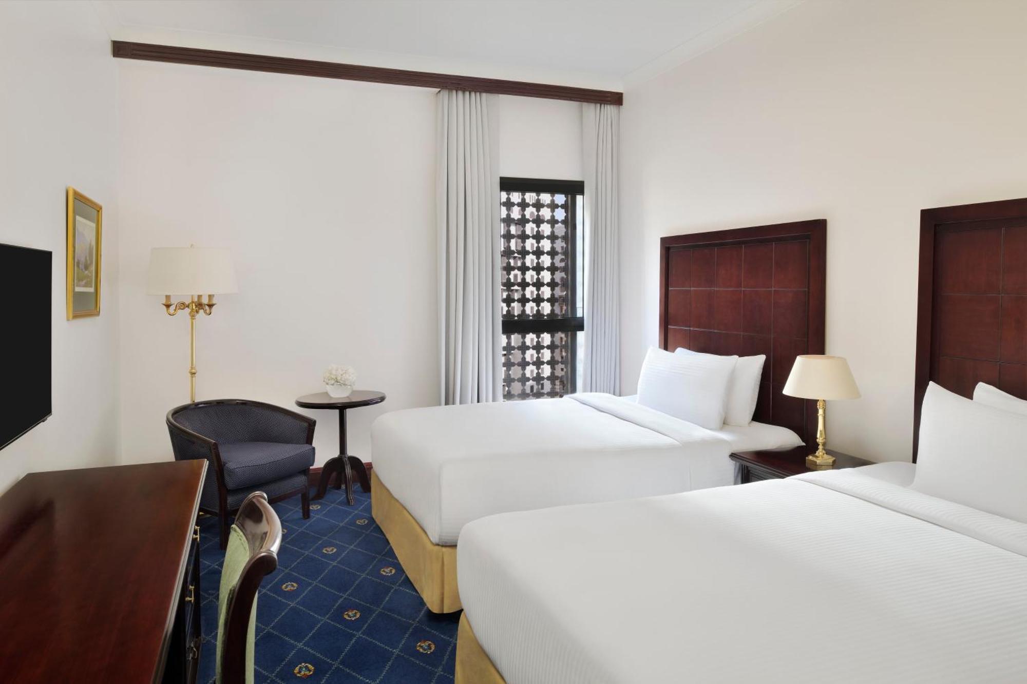 ダー アル ヒジラ インターコンチネンタル マディーナ エクステリア 写真 The photo shows a hotel room featuring two twin beds with white linens. There's a small table and chair in the corner, accompanied by a floor lamp. A wooden desk is positioned against one wall, and there is a window with sheer curtains, allowing natu