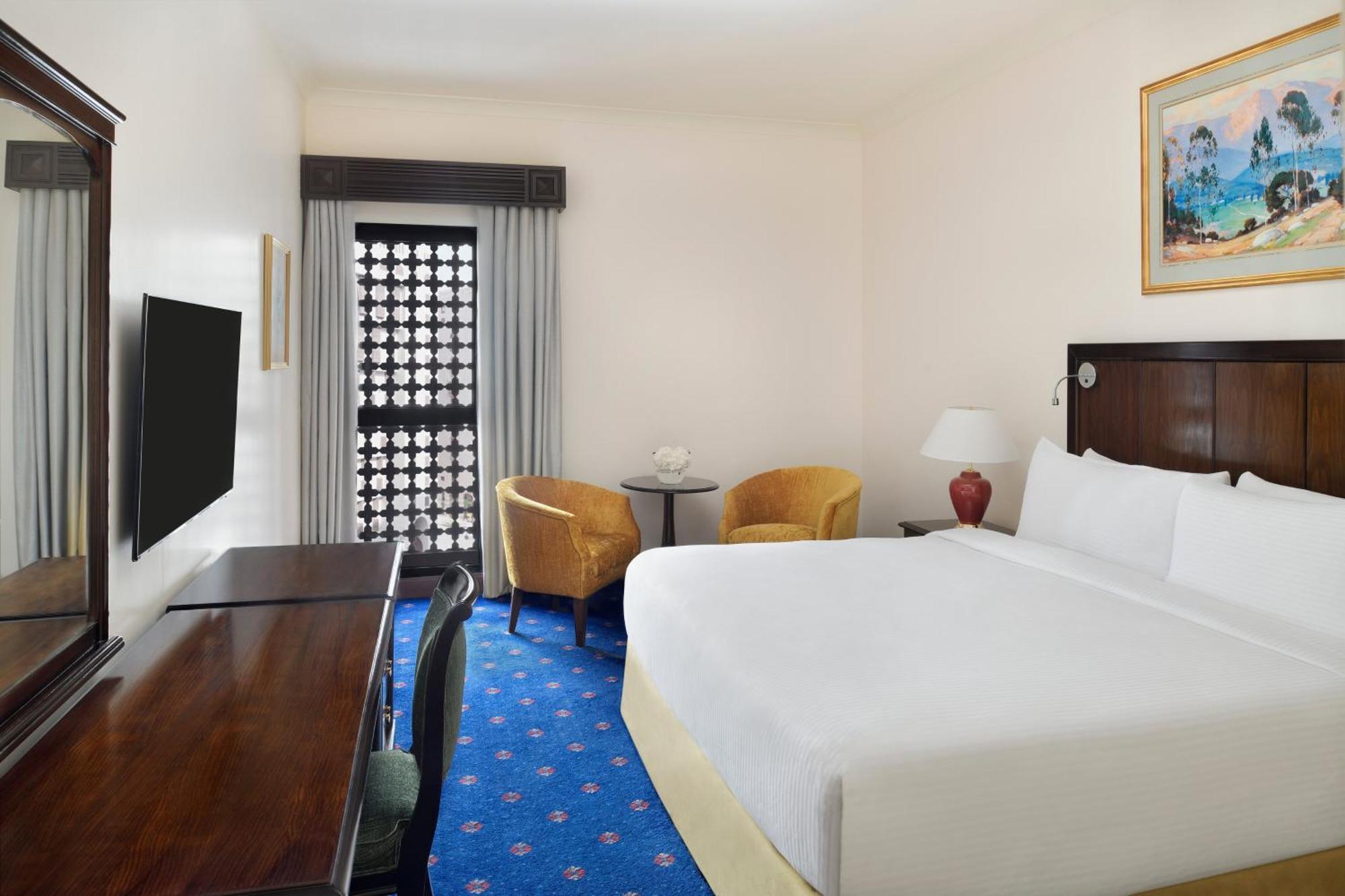 ダー アル ヒジラ インターコンチネンタル マディーナ エクステリア 写真 The photo shows a hotel room featuring a large bed with a white duvet and light-colored bed skirt. There is a desk with a chair on one side, and a modern television mounted on the wall. Two yellow armchairs and a small round table are positioned near