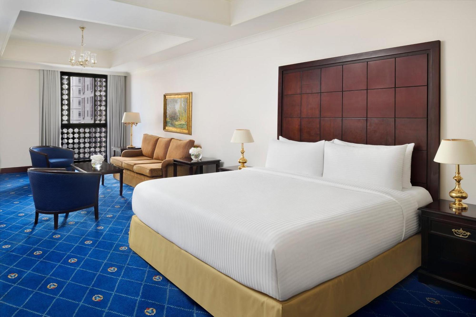 ダー アル ヒジラ インターコンチネンタル マディーナ エクステリア 写真 The photo shows a modern hotel room. It features a large bed with white linens and a light tan base. To one side, there's a comfortable seating area with a brown sofa and two blue chairs, along with a small table. A lamp is positioned on a side table