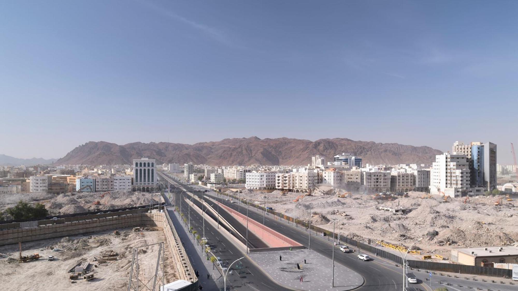 ダー アル ヒジラ インターコンチネンタル マディーナ エクステリア 写真 The photo shows a landscape featuring a wide road that runs through a city. On either side of the road, there are several modern buildings along with some under-construction sites. In the background, there are mountains that rise up, creating a rugge